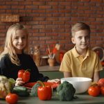 Positive little girl and boy sitting at table with various vegetables and herbs while preparing vegetarian meal in loft kitchen with brick wall